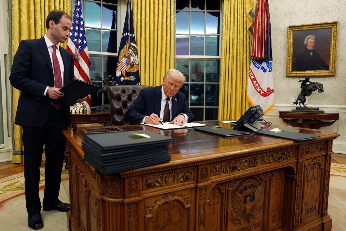 A man seated at a large desk signs a piece of paper while another man stands to his right.