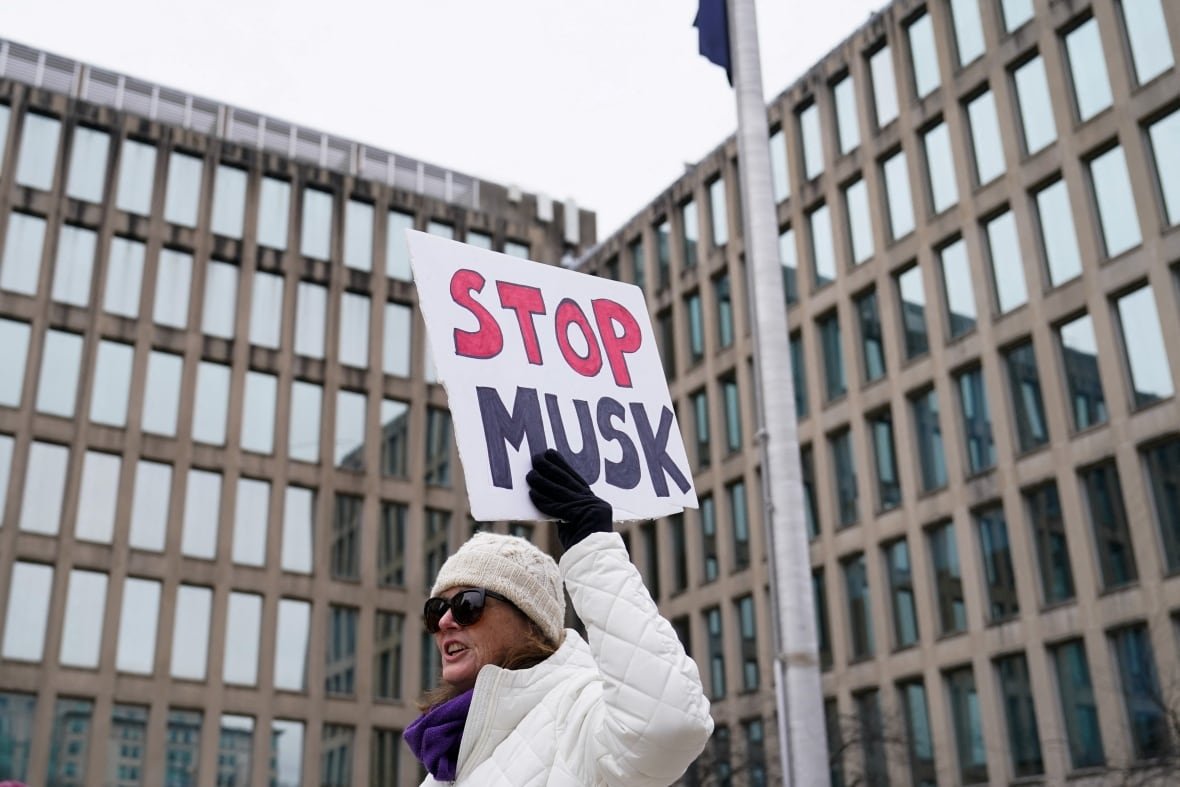 A woman wearing a toque and sunglasses holds a sign that says 'Stop Musk' outside a building.