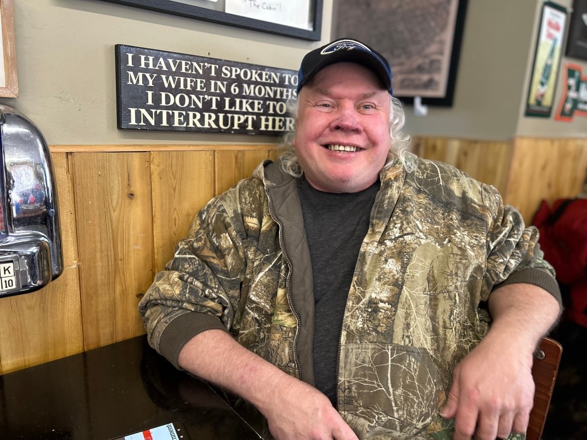 A man with a camouflage sweater and a ball cap smiles at the camera, while sitting at a table with signs on the wall and part of a jukebox player.