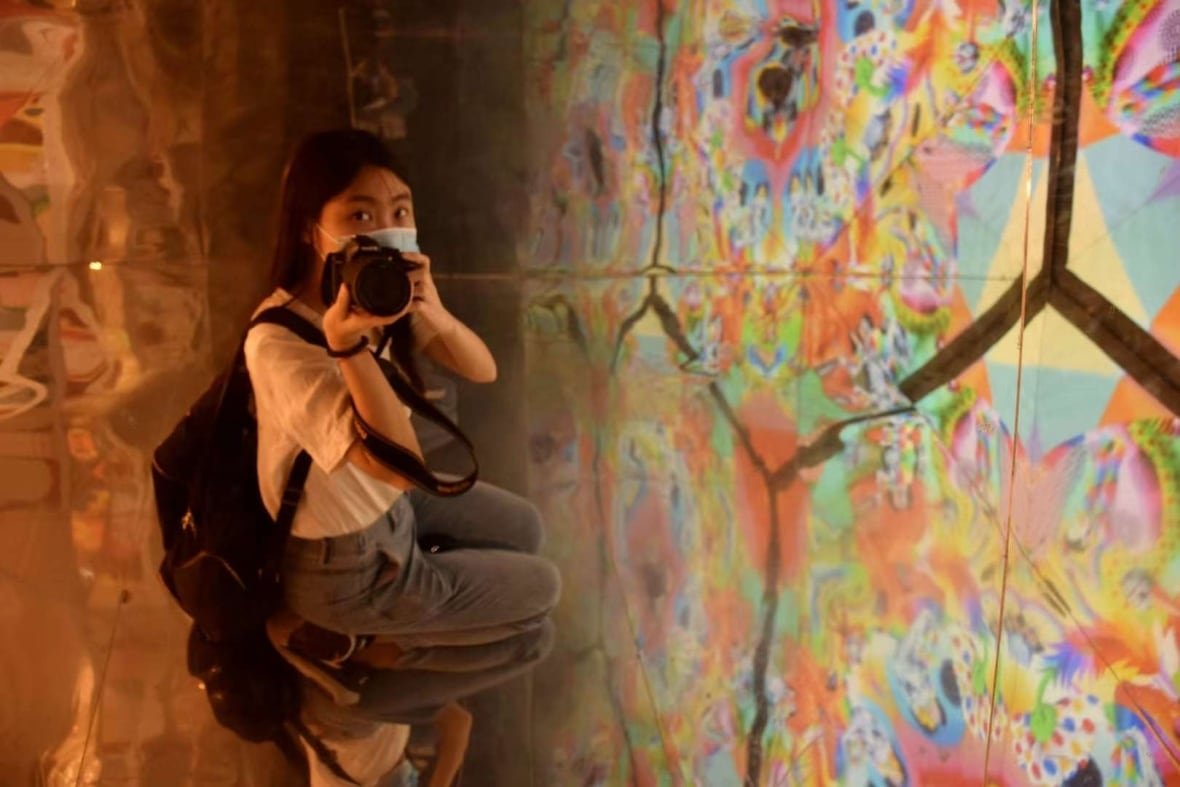 A woman wearing a surgical mask crouches down to take a photo of an art exhibit.