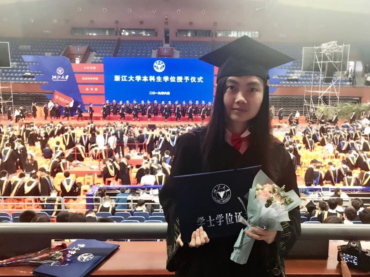 A smiling woman in a black graduation robe poses for a photo in an auditorium packed with other students wearing robes. She’s holding a bouquet and a certificate.