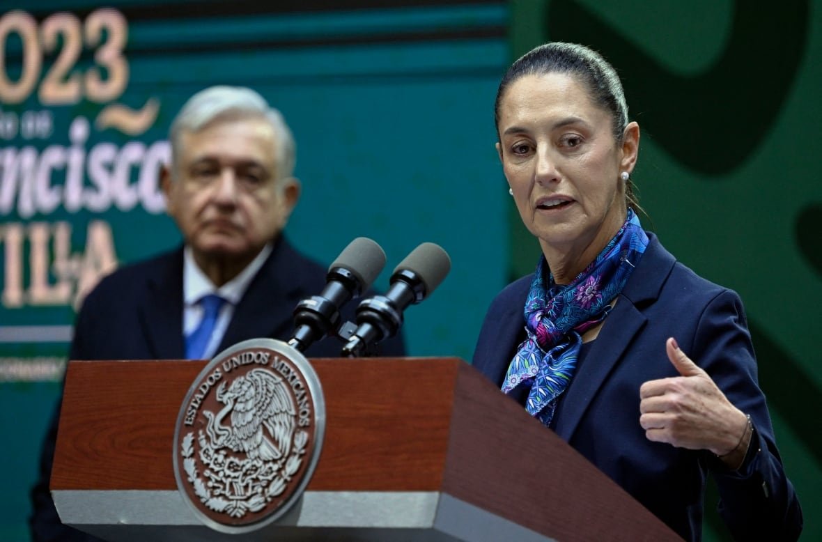 1246387807 A woman with her hair pulled back wearing a scarf speaks at a podium as an older man in a suit and tie looks on from the side.
