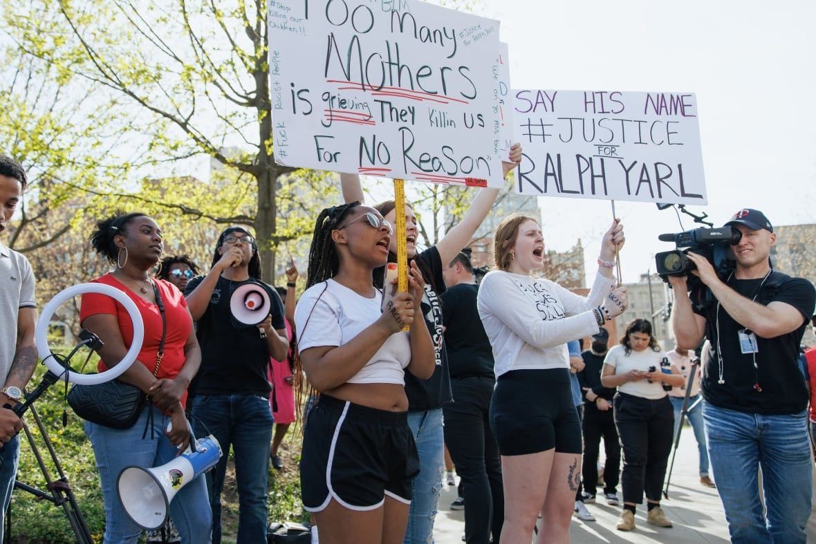 Several young looking people, men and women and dark-skinned and Caucasian, are shown at a demonstration. Some are holding up signs.