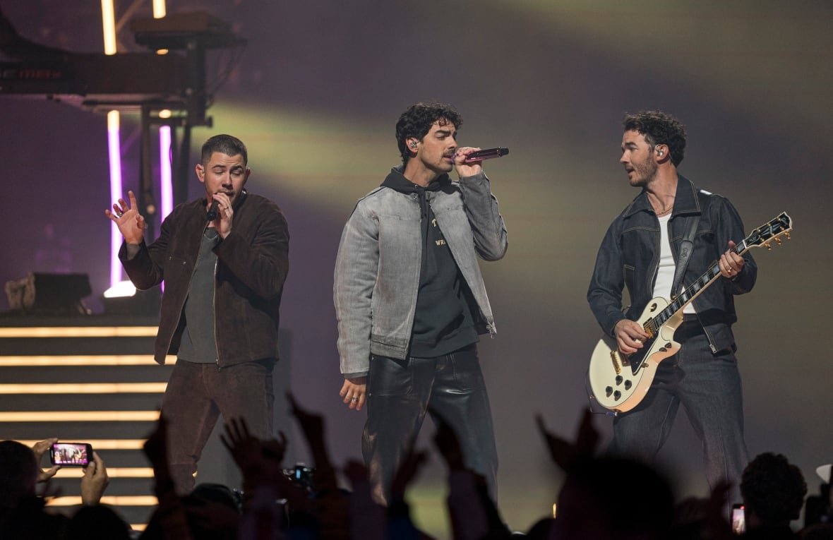 2184678711 Two men sing on stage while a third plays guitar