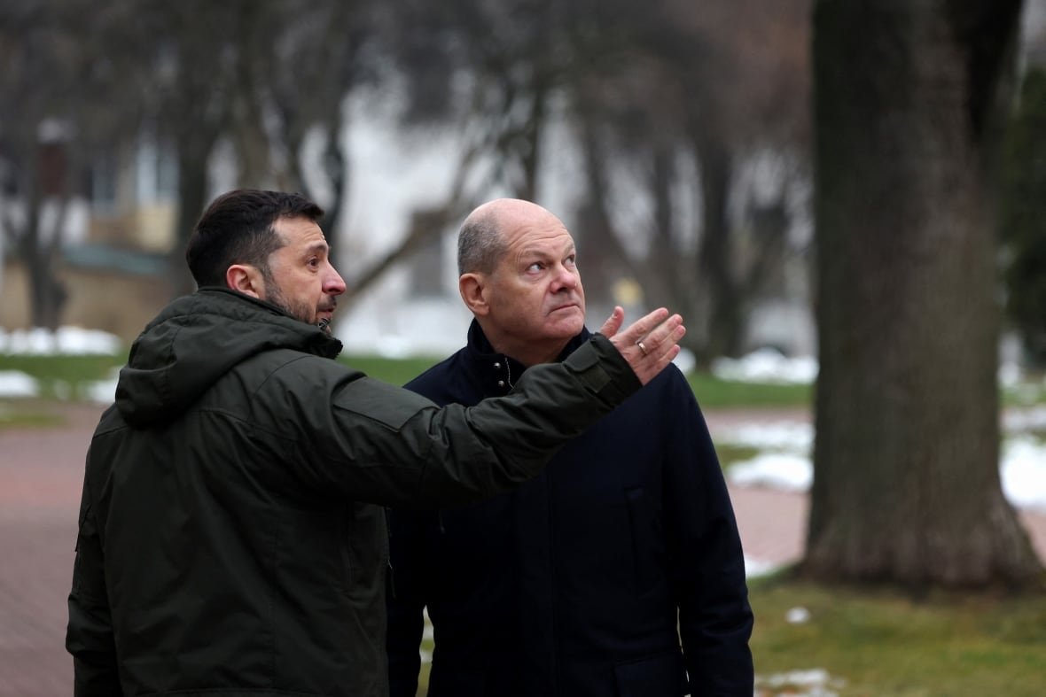 Two men, one older and balding and one younger, darkhaired and bearded, are shown outside talking and wearing coats.
