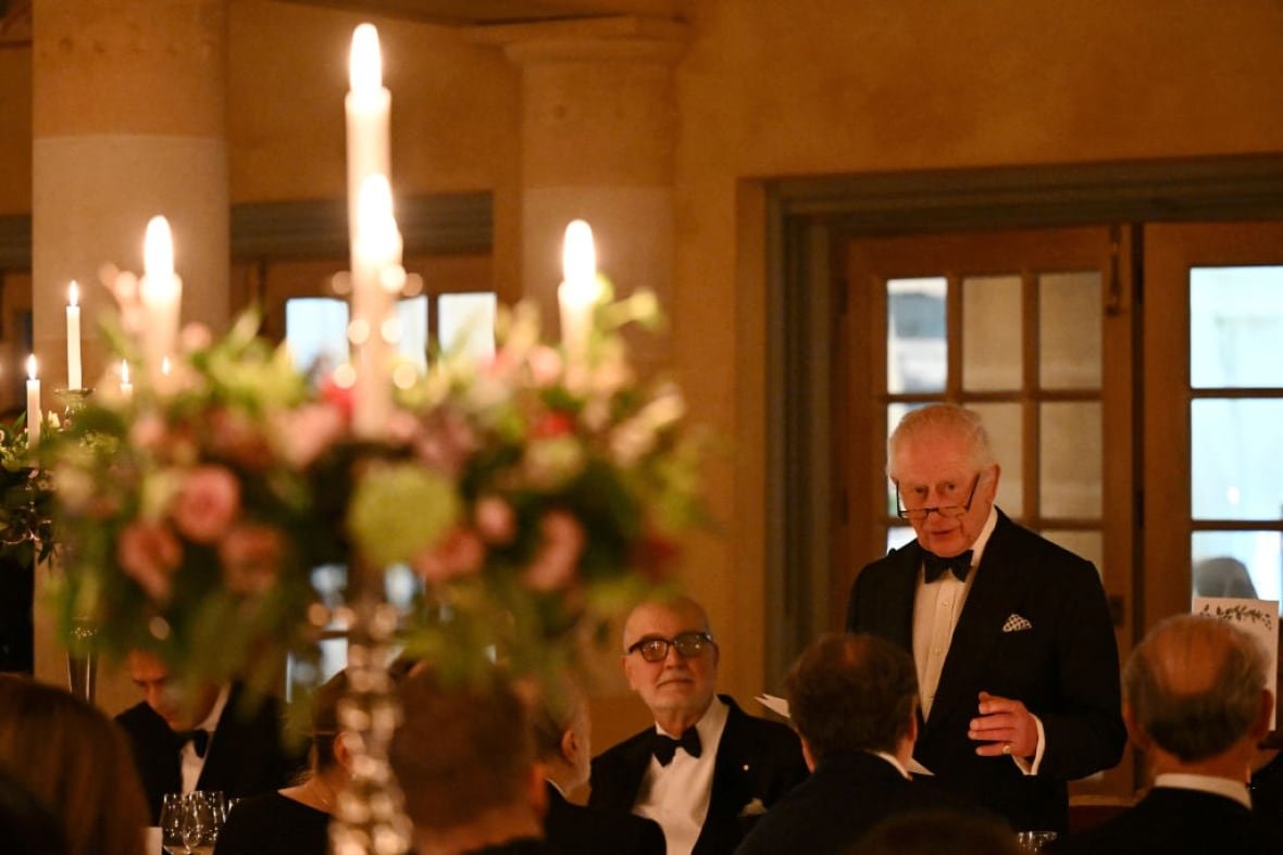 2197597002 A person holds papers as they speak to others seated at a formal dinner, with a large bouquet of flowers and candles in the foreground.