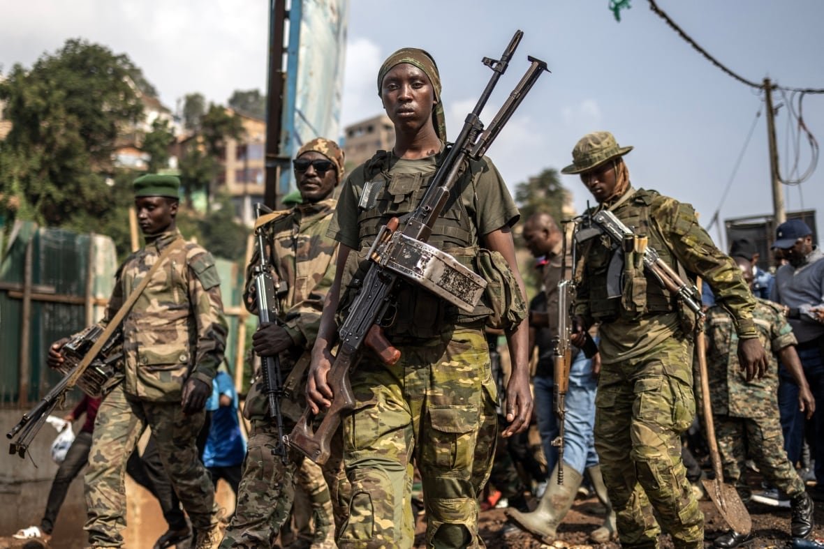 Several dark-complected man wearing camouflage and wearing long guns across their bodies are shown standing outdoors.