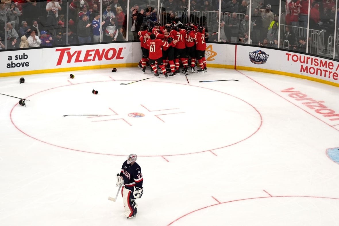 A hockey goaltender in a navy jersey skates away from a group of players celebrating in red jerseys.