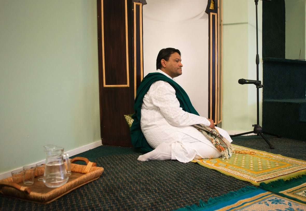598738212 A man in long white robes with a green fabric draped over his shoulder kneels on a carpeted floor near a pitcher of water.