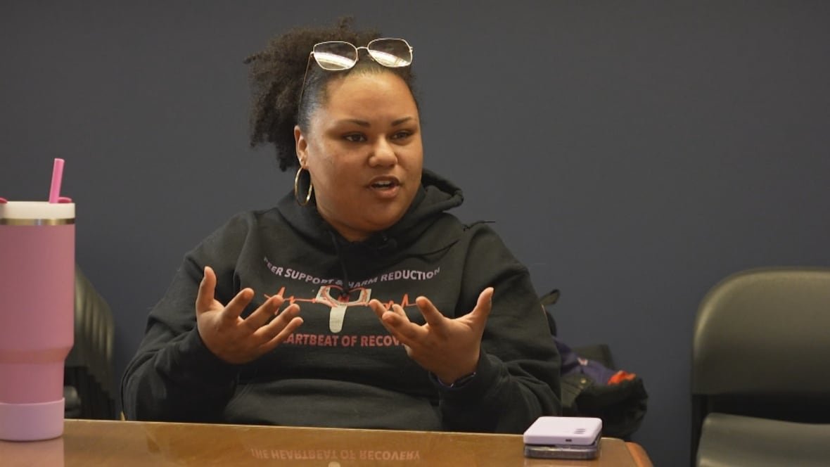 A woman in a hoodie is pictured sitting at a table speaking.
