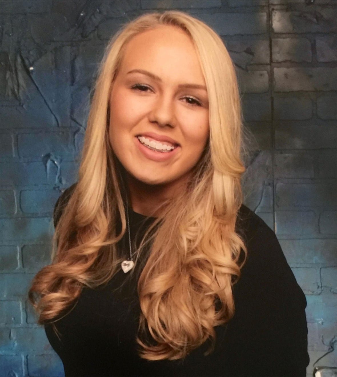 alexandra-capelouto A young blond girl smiles in front of a brick wall.