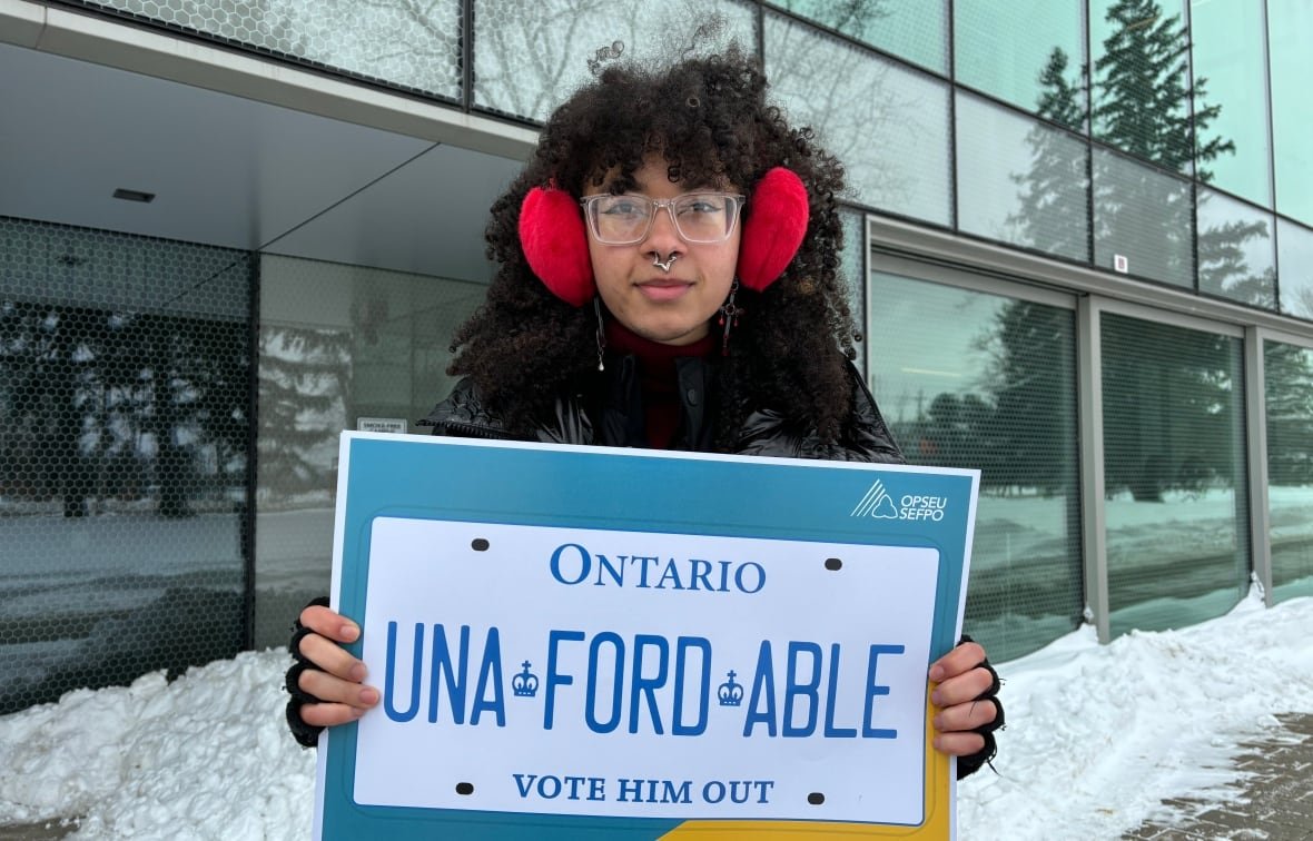 alexandria-catherine-davis-1 A person in a winter coat stands outside holding a sign that looks like a licence plate and says "Una-FORD-able."