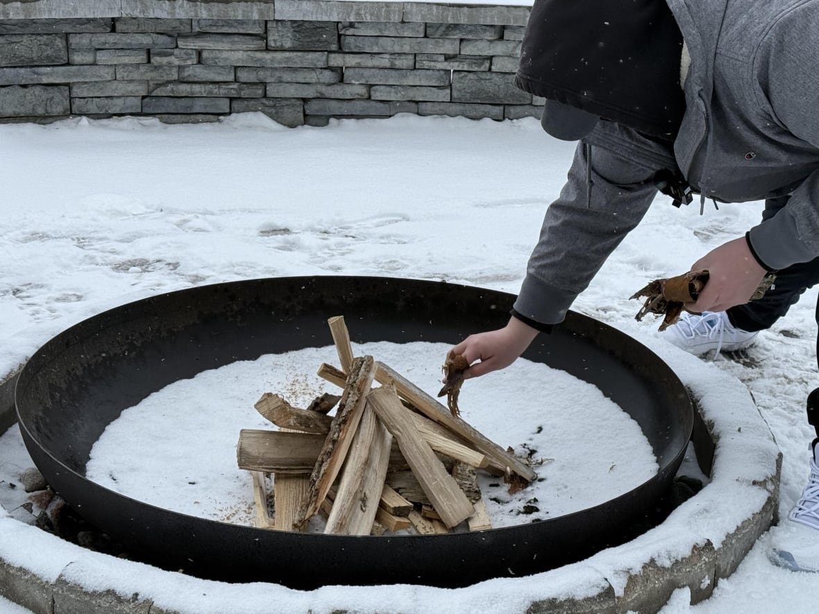 ann-s-eye-february-16-2025-1 A man wearing a grey winter jacket and a black hood bends over an outdoor fire pit and adds wood shavings to pieces of wood.