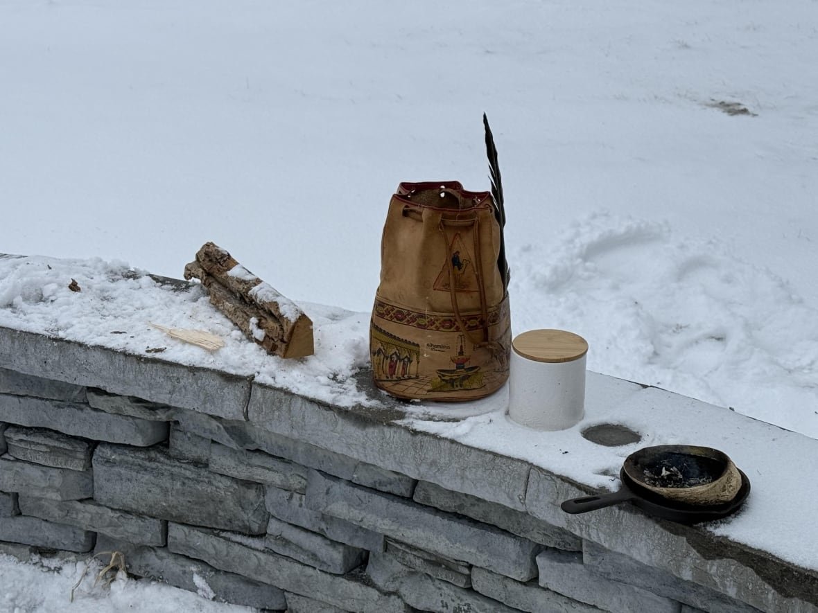 ann-s-eye-february-16-2025-2 A brown bag with a feather on the side sits on a short, snow-covered stone wall in between a log of wood and a small white jar.