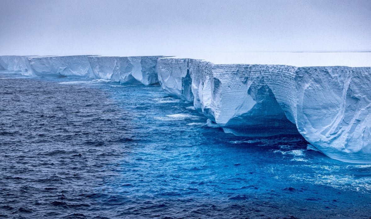 A giant iceberg is seen from the side, floating in the ocean. There are big pits and dips seen in the side of the iceberg, but the top is flat.