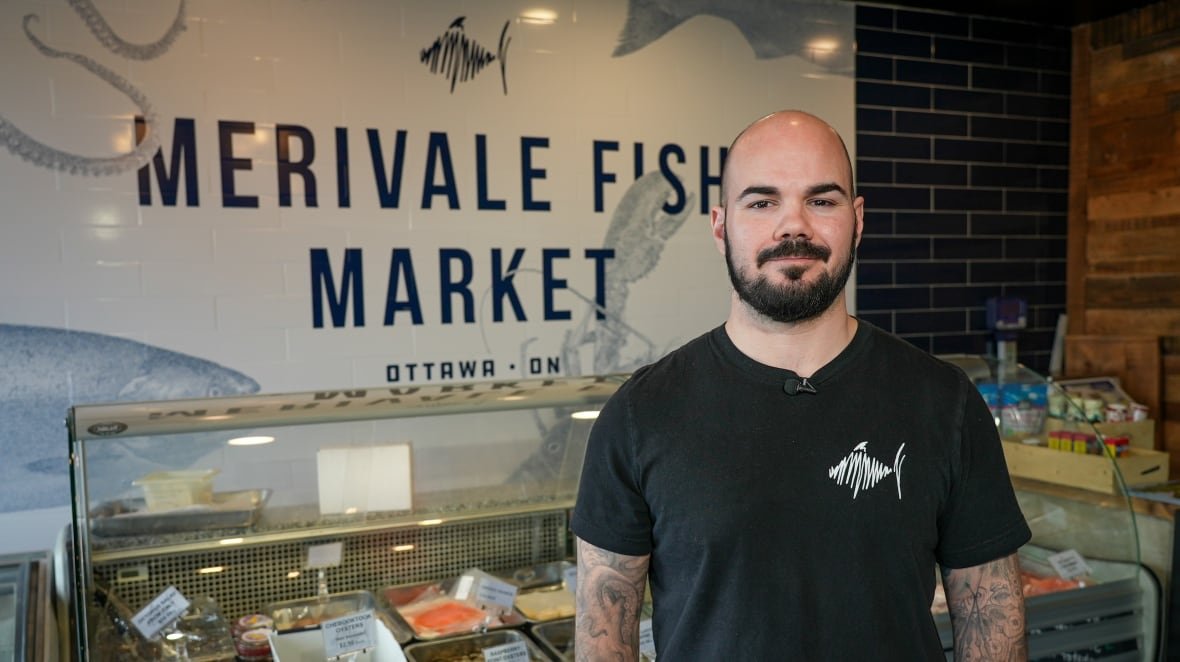 A bald man with a beard stands in front of a fish cooler and a sign that reads "Merivale Fish Market."