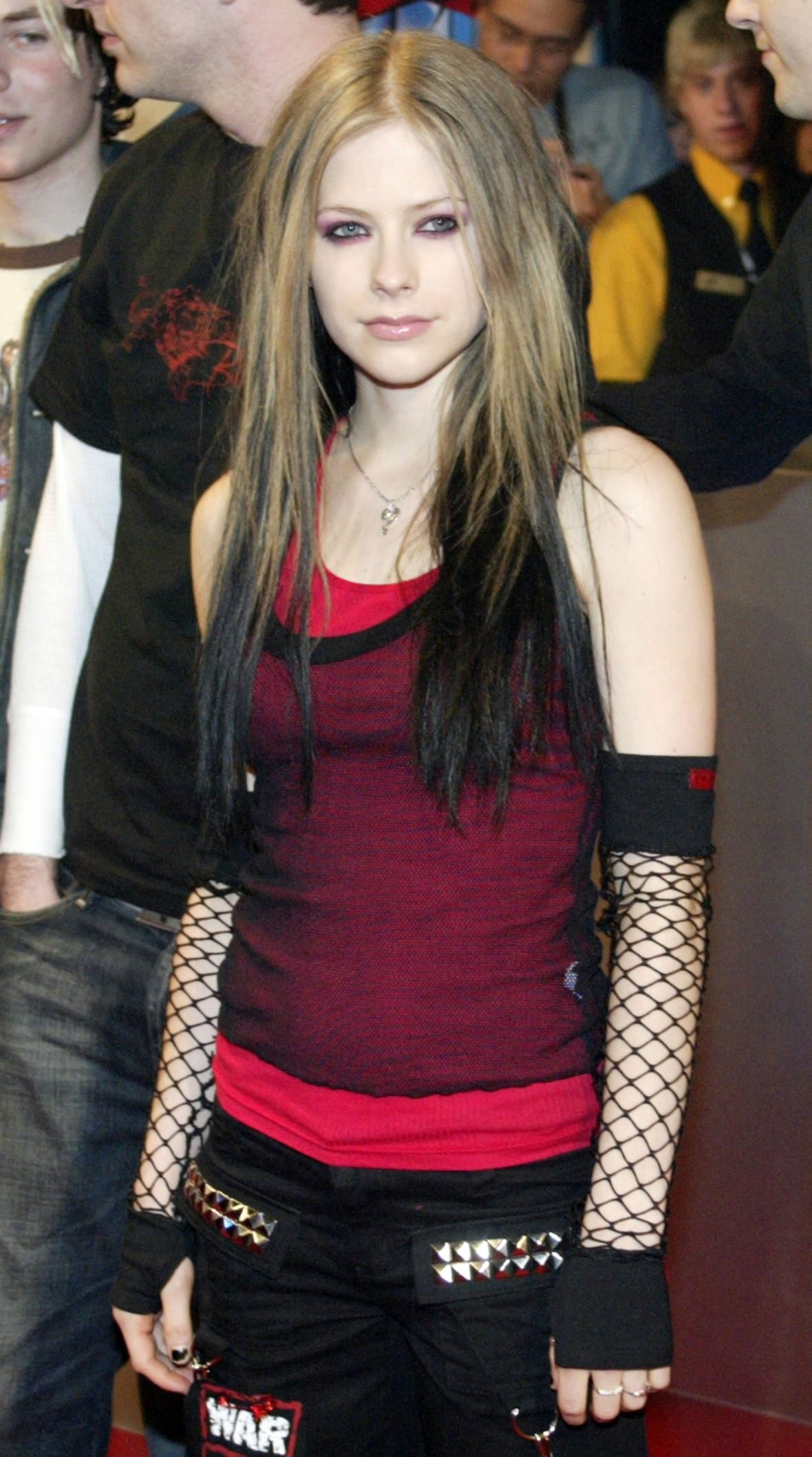 Avril Lavigne on the red carpet wearing black pants, a red tank top with a sheer black tank top over it, and long fishnet gloves.