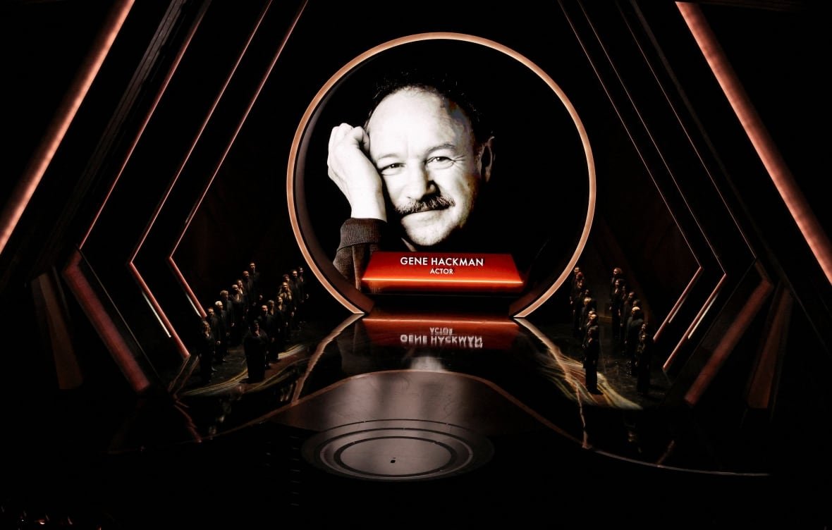 A black and white image of a smiling man with a moustache is projected on a darkened stage in a theatre.