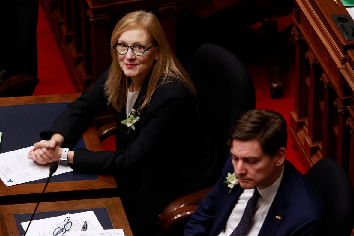 bc-throne-speech-20250218-1 A red-haired woman in a black, formal top sits with her hands clasped in front of her on a wooden table. A man with short brown hair is seen out of focus to her right.