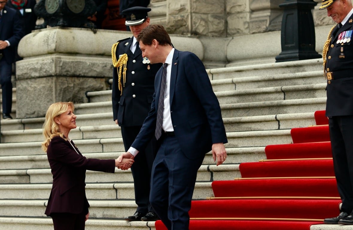 bc-throne-speech-20250218 A tall man in a suit stands outdoors on a set of red-carpeted steps and reaches down to shake hands with a blond-haired woman in a black business suit.