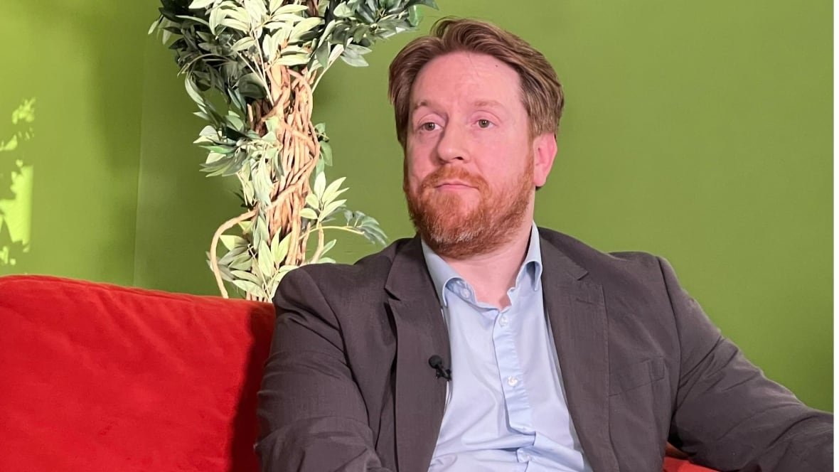 ben-wickstrom A man in a suit jacket is seated on a red couch in front of a green wall with a plant behind his shoulder.