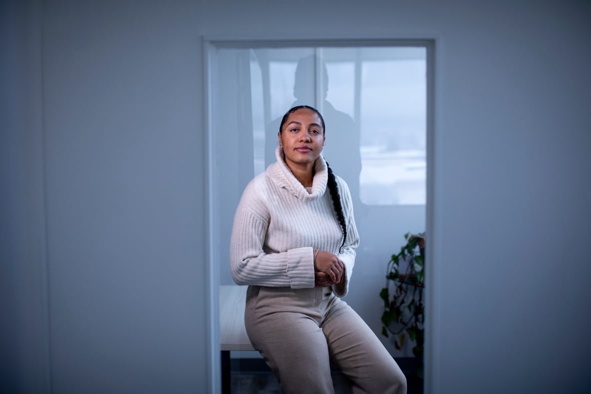 A light-skinned Black woman wears a white turtleneck sweater and looks straight at the camera.