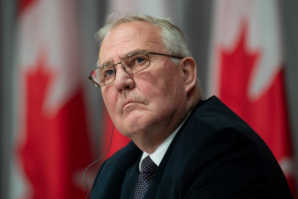An older man in a suit looks to his left. There are many Canadian flags behind him.