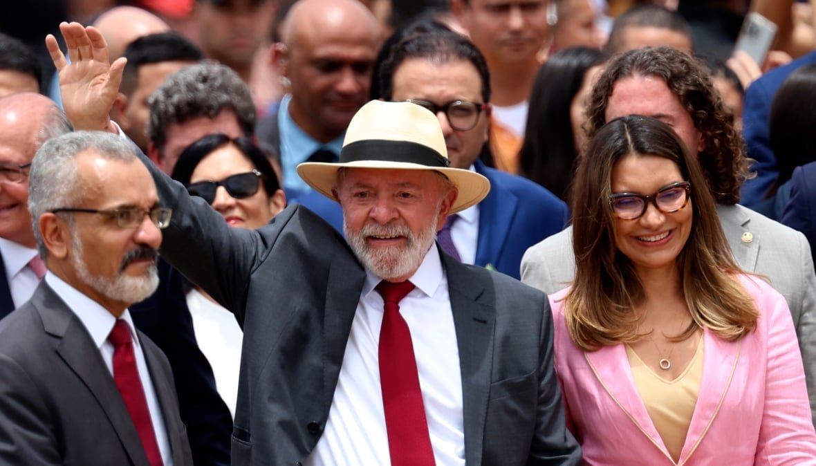 A man waves to a crowd while standing next to a woman in a pink blazer. A number of people are standing behind him.