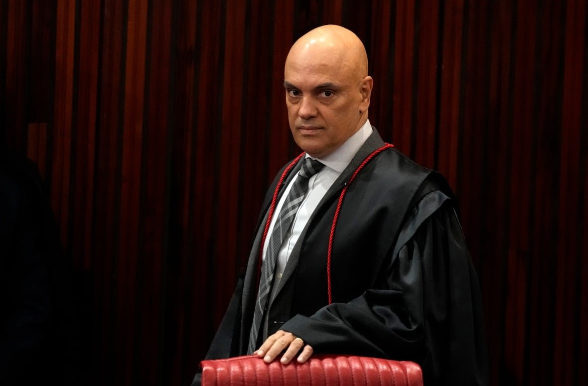 A man in a judge robe stands in front of a red-paneled backdrop, looking at the camera with his hand on the back of a red chair.