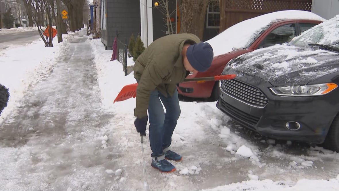 bruce-rands A man holding a shovel and a golf club tries to break up ice in his driveway. There are two cars parked nearby, and the man wears a green jacket, a blue toque, and jeans.
