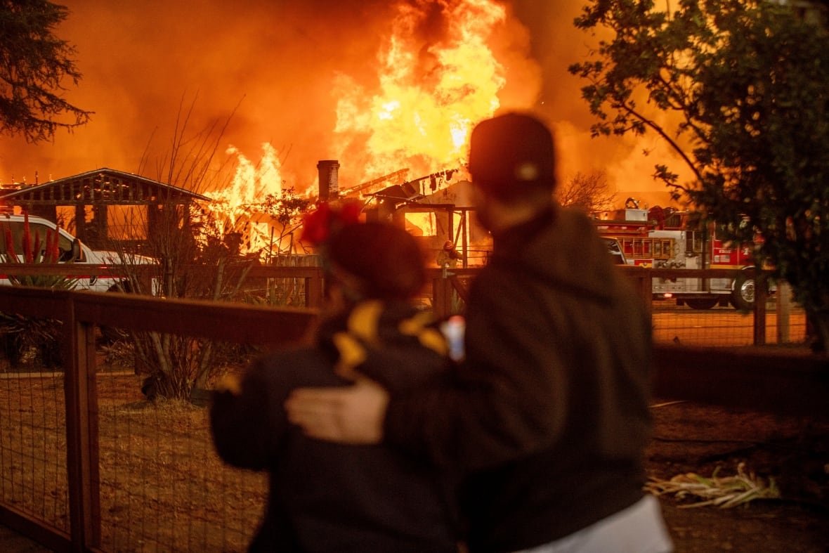 california-wildfires-altadena-residents-1 Two people, with their backs turned, faces a small house engulfed in flames.