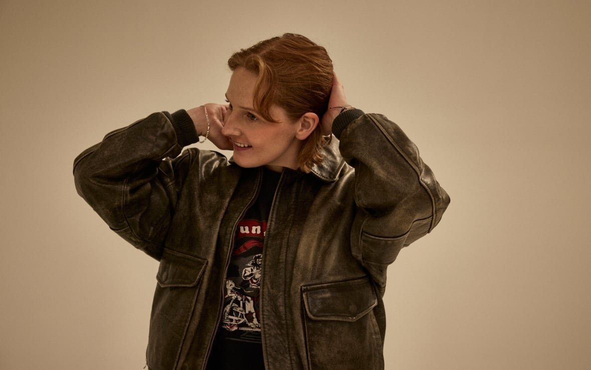 A young woman wearing a leather jacket smiles as she runs her hands through her short, red hair.