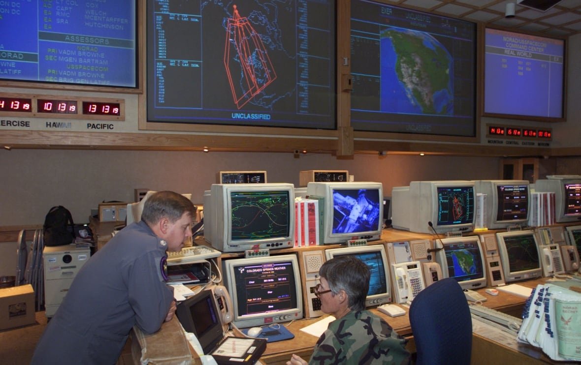 Two men in military uniforms work amid a bank of computer terminals, facing a wall of large screen displays.
