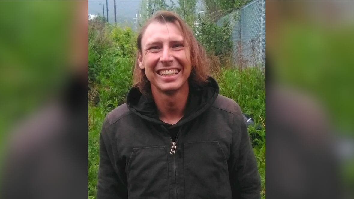 A young man smiles while standing outside in front of some green vegetation. He has chin-length hair and is wearing a black hoodie and zip-up jacket.