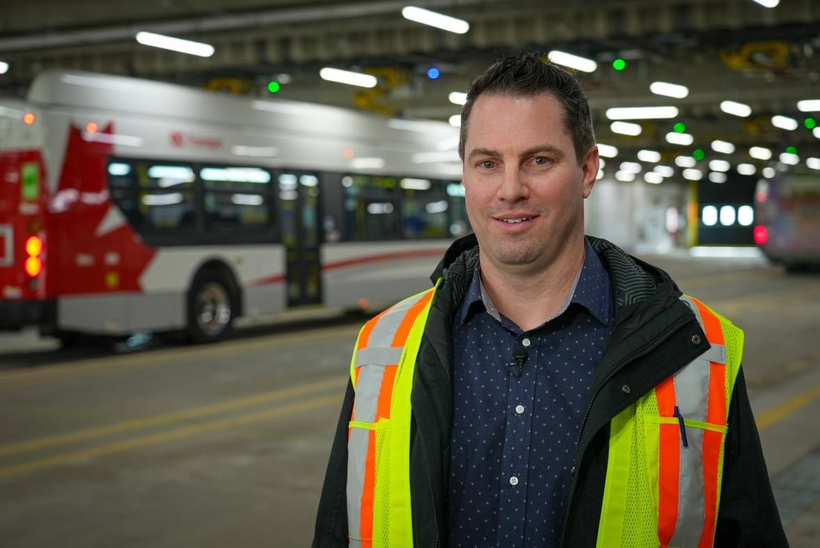 daniel-villeneuve-manager-zero-emission-bus-program-jan-30-2025-1 A man in a florescent safety vest stands in front of a bus in a nearly empty garage