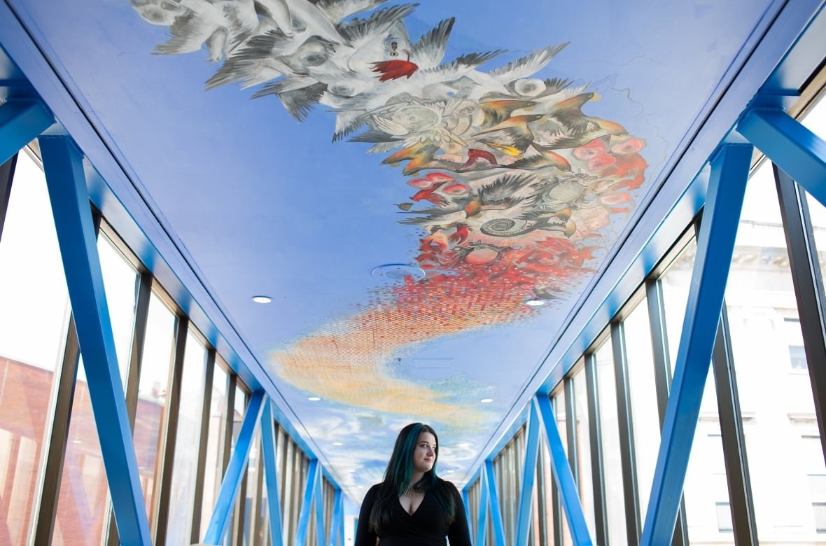 deanna-musgrave-nest A woman standing in a pedway with a large mural above her, showing a cluster of birds and an optical illusion of sorts.