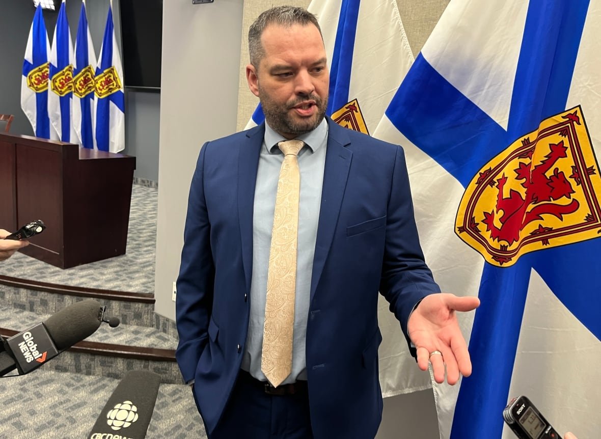 A man in a suit and tie stands at a row of microphones with Nova Scotia flags behind him.