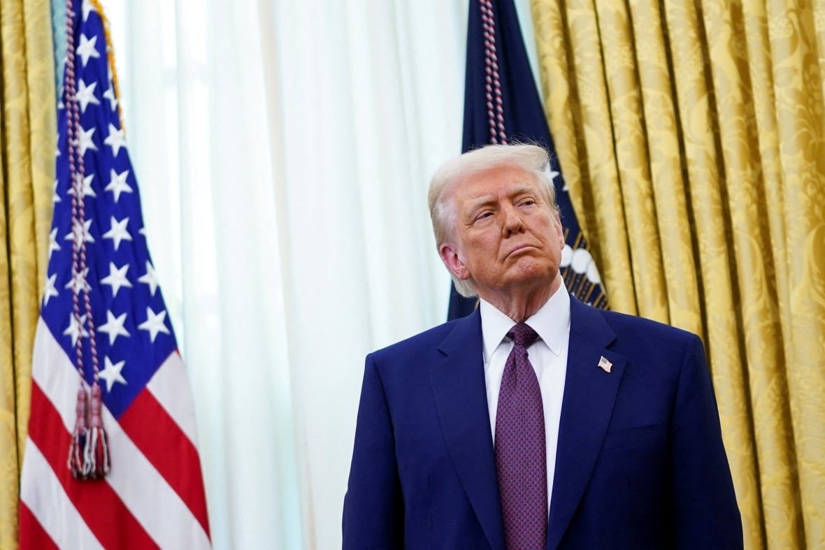 donald-trump-1 A man in a navy suit with a purple tie stands in an office with American flags behind him.