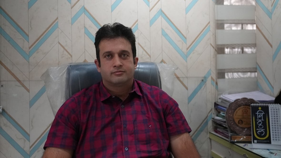 A man with black hair and brown eyes is sitting in a chair and looking at the camera with a neutral expression. He is wearing a red and blue plaid shirt.