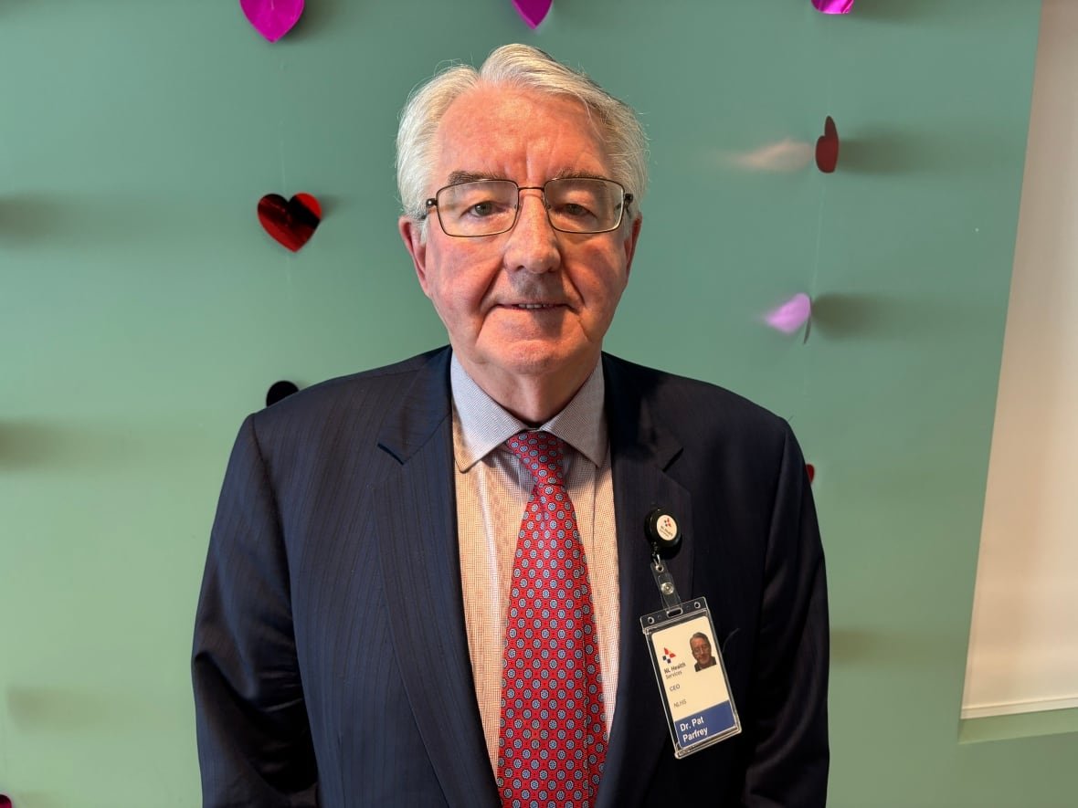 A smiling man wearing a suit stands against a turquoise wall. The wall has paper hearts on it as a decoration for Valentine's Day.