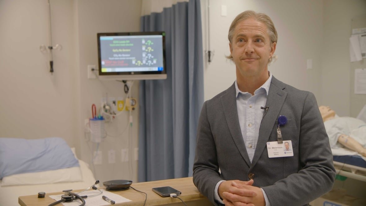 A man wearing a blue collared shirt and a grey blazer, with an identification tag pinned to his breast pocket, standing in a hospital room, with a medical manikin in the bed behind him.