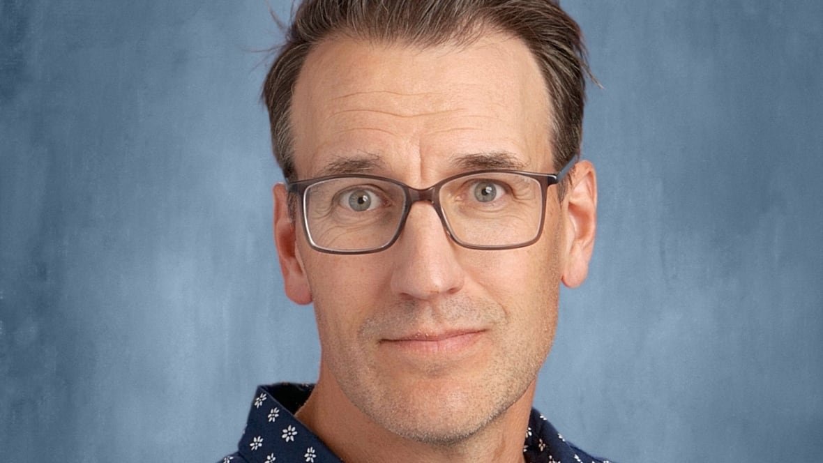 Drew Van Allen is a white man with blue eyes and brown hair, he wears glasses and sits against a blue backdrop.
