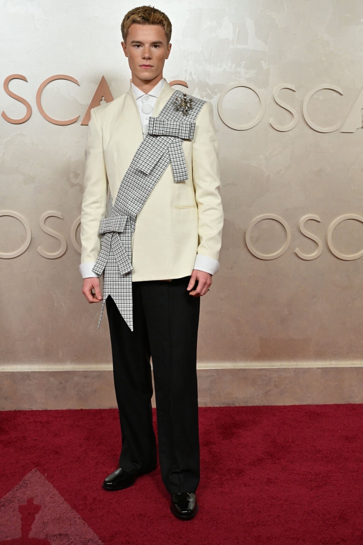 edvin-ryding-attends-the-97th-annual-academy-awards Edvin Ryding on the red carpet wearing a cream-coloured suit jacket, black pants and a grey and white check sash and oversized silver floral brooch.