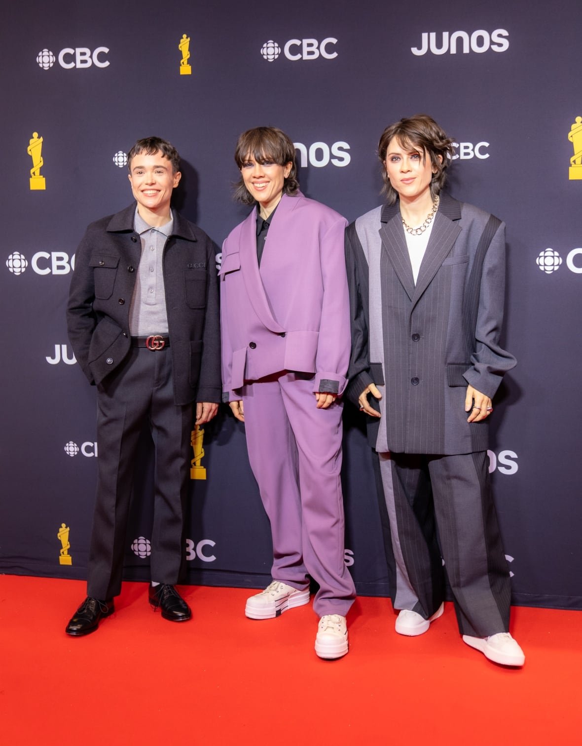 Elliot Page, Tegan Quin and Sara Quin on the red capet. Elliot is wearing grey dress pants, grey shirt and grey jacket with a Gucci belt. Sara is wearing an oversized purple suit. Tegan is wearing an oversized grey suit.