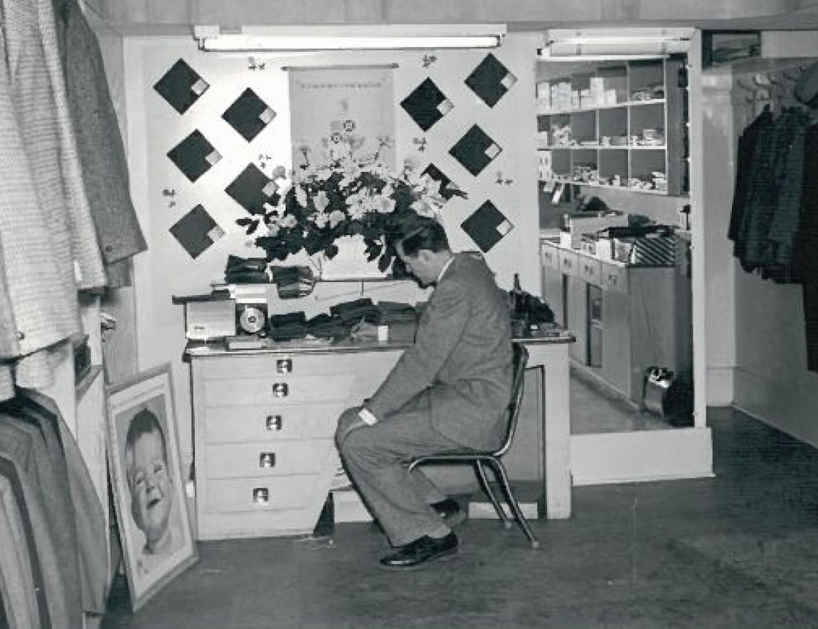 elwood-flynn A black and white photo of a man sitting a desk in a men's clothing store.