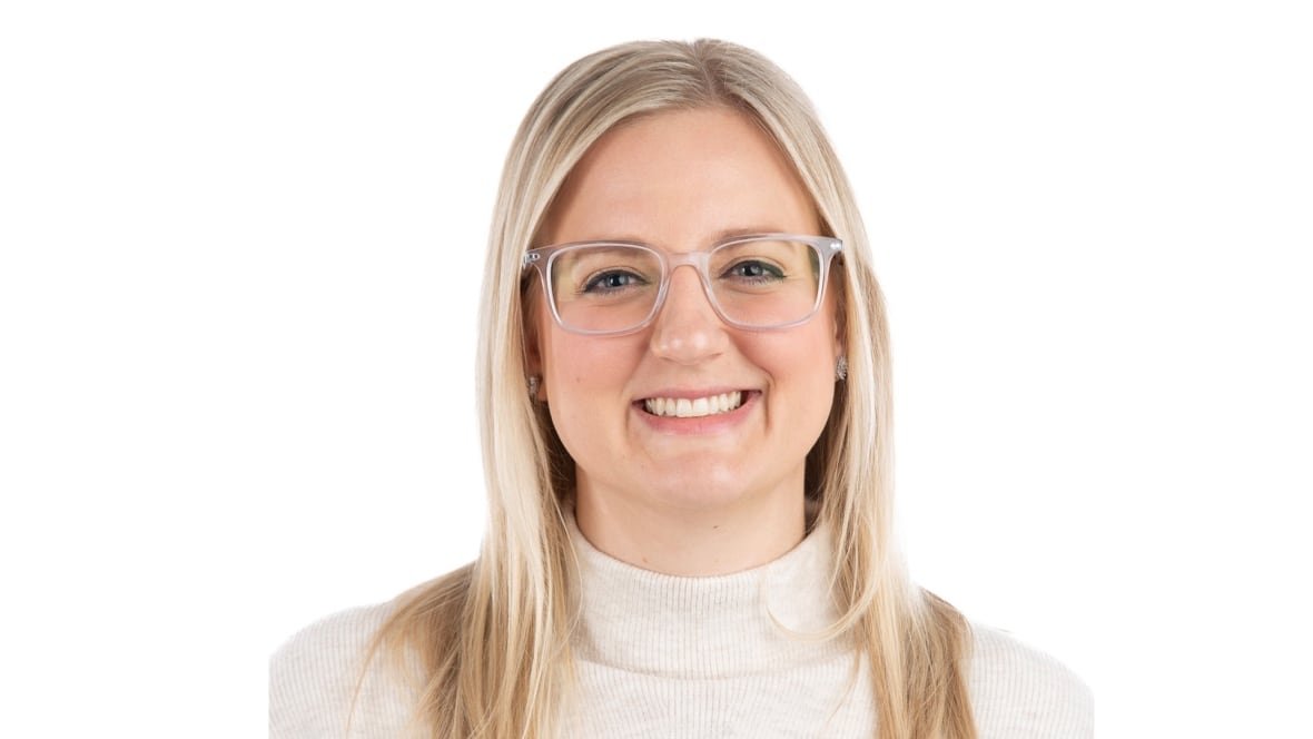 Emily has blonde hair and blue eyes, she smiles while wearing dark rimmed glasses and a beige mock neck sweater.