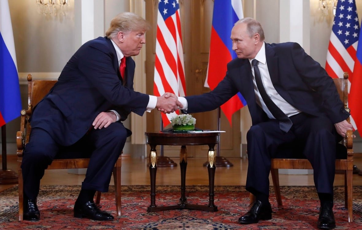 Two men shake hands across a table, with flags in the background.