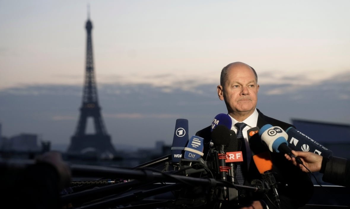 A man stands in front of a bunch of microphones, all with different news labels on them. He is standing outside at twilight and in the distance is the blurry silhouette of the Eiffel Tower.