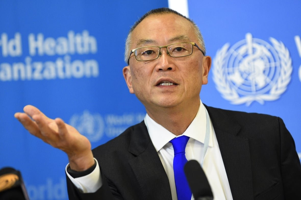 Man in a dark suit with a blue tie wearing glasses and holding his right palm up.