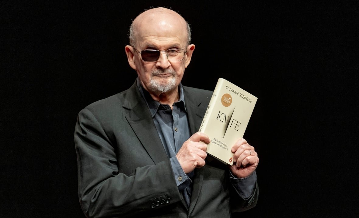 A man holds up a book in front of a black backdrop. He is wearing a suit.